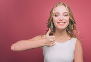 Cheerful and attractive young person is holding her thumb up and smiling. She is very cheerful. Isolated on pink background.